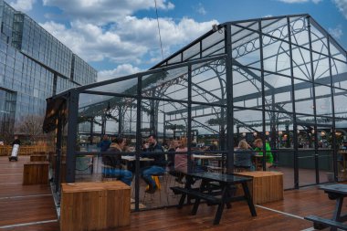 Londra 'da bir çatı katında cam kaplı bir yemek alanında metal kemerler, ahşap koltuklar ve şenlikli dekorasyonlar bulunur. İnsanlar parçalı bulutlu bir gökyüzünün altında yemek yerler..