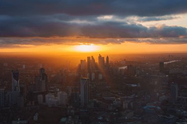 Görüntü Londra 'da gün batımını gösteriyor. Kanarya Rıhtımı gökdelenleri, ufukta altın bir parıltı, kara bulutlar ve Thames Nehri şehrin içinde dolanıyor..