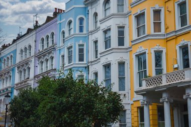 Notting Hill, Londra 'da bir sıra pastel renkli Viktorya dönemi evleri. Süslü detaylar ve bulutlu gökyüzünün altındaki yemyeşil bir ağaç. Şehir mimarisini ön plana çıkarıyor..