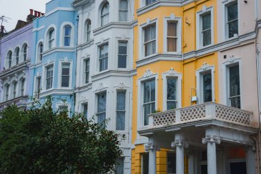 Londra 'daki Notting Hill' deki renkli Victoria tarzı teraslarda pastel renkler ve süslü detaylar yer alıyor. Ön planda yeşillik ve bulutlu bir gökyüzü sahneyi tamamlıyor.