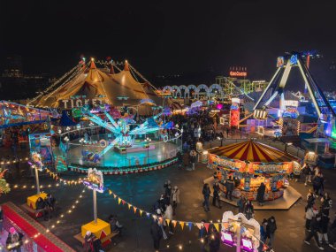 A bustling Christmas fair in London features a large tent marked 'TICKETS', colorful rides like a carousel and roller coaster, and a lively crowd at night. clipart