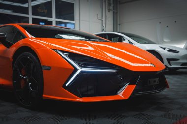 Modern bir galeride turuncu bir Lamborghini Revuelto sergilenmektedir. Arabanın açısal farları ve aerodinamik tasarımı arka planda beyaz bir araba ile vurgulandı.