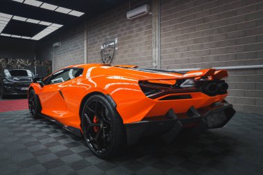 Canlı turuncu bir Lamborghini Revuelto, duvarları tuğlalı ve arkası kareli bir sergi salonunda sergileniyor..