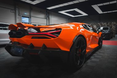 Turuncu bir Lamborghini Revuelto, keskin çizgiler, özel arka ışıklar ve kareli zeminde çift egzozla iyi aydınlatılmış bir sergi salonunda sergilenir..