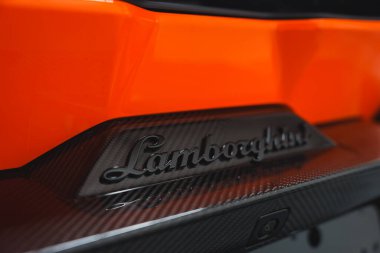 Görüntü Lamborghini Revuelto 'nun arka bölümünü gösteriyor. Karbon fiberine karşı hazırlanmış ambleme odaklanıyor. Canlı turuncu kaportayla..