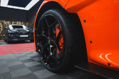 Görüntü Lamborghini Revuelto 'nun arka tekerleğini ve fren kalibresini yakalıyor. Canlı turuncu araba kapalı bir yerde desenli bir zemine göre dizilmiş..