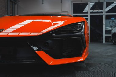 Görüntü Lamborghini Revuelto 'nun ön tarafının yakın çekimini gösteriyor. Açısal tasarımını, canlı turuncu rengini ve kapalı alandaki eşsiz farlarını vurguluyor..