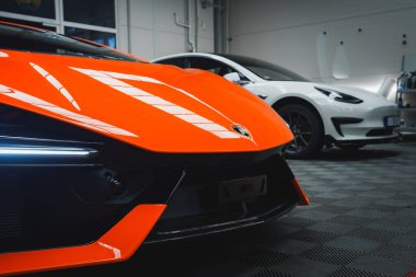 Canlı turuncu bir Lamborghini Revuelto parlak ön tasarımı ve ayırt edici farlarıyla sergilenir. Arka planda beyaz bir Tesla var..