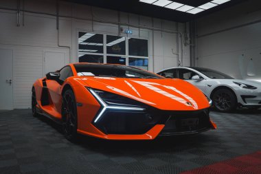 Turuncu bir Lamborghini Revuelto kapalı bir yere park edilmiş, açısal farlarını ve aerodinamik vücudunu sergiliyor. Yanında beyaz bir araba var..