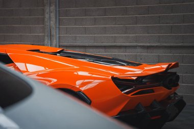 Görüntü turuncu bir Lamborghini Revuelto 'nun arka kısmını yakalıyor, parlak tasarımını, keskin çizgilerini ve gri tuğla bir duvara çarpan ayırt edici stop lambalarını vurguluyor..