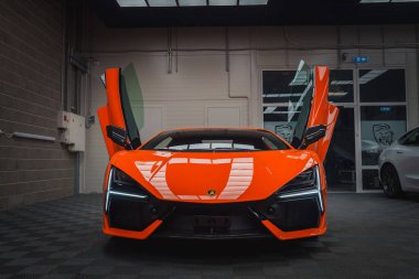 Turuncu bir Lamborghini Revuelto kapalı mekanda sergileniyor. Makas kapıları açık, parlak tasarımı sergileniyor. Ayarda kareli bir zemin ve tepe ışıkları yer alıyor.
