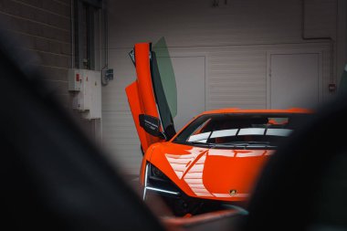 Canlı turuncu bir Lamborghini Revuelto makas kapısı açık bir şekilde kapalı mekanda sergilenir. Ayarlar arasında bir tuğla duvar ve arka planda elektrik panelleri yer alıyor..
