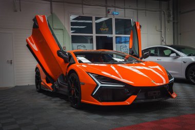 Turuncu bir Lamborghini Revuelto kapalı bir yere park edilmiş, makaslı kapılar açık. Yansımalar, yakınında beyaz bir araba bulunan tasarımını vurgular..