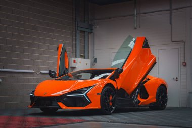 Turuncu bir Lamborghini Revuelto kapalı mekanda makas kapıları açık bir şekilde sergileniyor. Parlak tasarımını endüstriyel duvarlara ve fayanslı zemine vurguluyor..