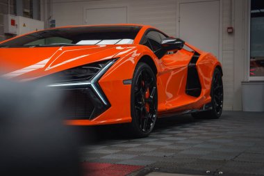 Turuncu bir Lamborghini Revuelto kapalı alanda sergileniyor, parlak tasarımı, modern LED farları ve fayanslı döşeme üzerindeki parlak kaplaması vurgulanıyor.