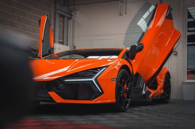 Turuncu bir Lamborghini Revuelto, makas kapıları açık, açısal farlarını ve aerodinamik tasarımını beton bir duvara karşı sergiliyor..