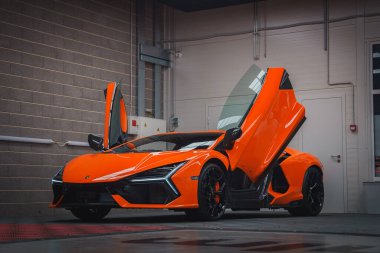 Turuncu bir Lamborghini Revuelto kapalı mekanda sergileniyor, makas kapıları açık, beton bir duvara ve endüstriyel elementlere karşı dizayn edilmiş,.