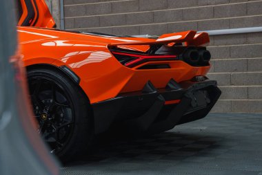 Turuncu bir Lamborghini Revuelto kapalı bir yerde gri tuğladan bir duvara park edilmiş, parlak tasarımı, ayırt edici arka lambaları ve çift egzozları sergileniyor..