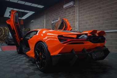 Kapıları açık olan turuncu bir Lamborghini Revuelto kapalı bir yerde kareli zeminde sergileniyor, aerodinamik çizgilerini ve çift egzozlarını vurguluyor..