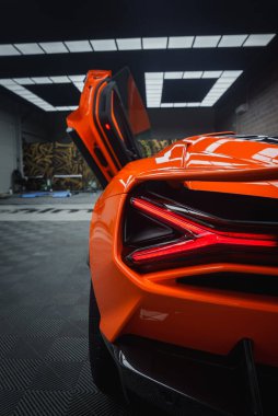 Görüntü, turuncu bir Lamborghini Revuelto 'nun arka bölümünü, aerodinamik tasarımını, ayırt edici arka lambalarını ve makas kapısını,.