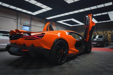 Turuncu bir Lamborghini Revuelto, aerodinamik tasarımını sergileyen makas kapıları açık bir şekilde kapalı mekanda sergilenir. Ayarlar modern bir tavan ve kareli zemin içeriyor.