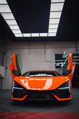 Turuncu bir Lamborghini Revuelto kapalı mekanda, makas kapıları açık, modern bir tavanın altında parlak yüzeyine yansıyan ışık panelleriyle sergilenir..