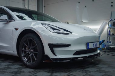 Beyaz bir Tesla Model 3 kapalı bir yerde kareli bir zeminde, araba bakım araçlarıyla çevrili. Işıklandırma parlak tasarımını ve parlak sonunu vurguluyor..