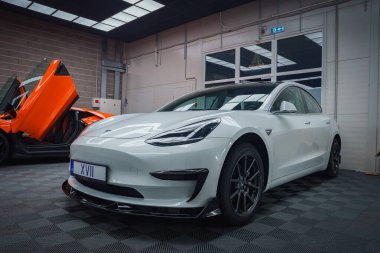 Plakası XVII olan beyaz bir Tesla Model 3 kapalı bir yerde, kareli bir zeminde park halinde. Açık makas kapılı turuncu bir Lamborghini kısmen görülebilir..