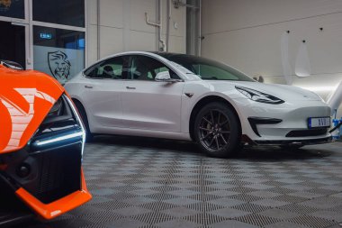 Beyaz bir Tesla Model 3, ön planda kısmen görülebilen turuncu renkli Lamborghini Revuelto ile desenli bir zemine park edilmiştir..