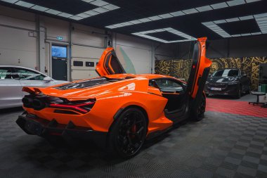 Modern bir garajda açık makas kapıları olan canlı turuncu bir Lamborghini Revuelto. Damalı zemin ve diğer arabalar dinamik bir zemin oluşturur.