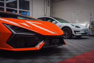Parlak turuncu bir Lamborghini Revuelto, beyaz bir Tesla Model 3 'ün yanına, kapalı bir zemine park edilmiştir. Ayarlar araçları ve ekipmanları içerir.