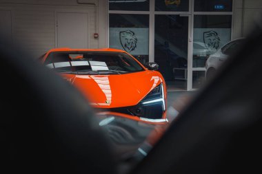 A vibrant orange Lamborghini Revuelto is showcased indoors, featuring LED headlights and emblem. Glass doors with lion logos are visible in the background. clipart