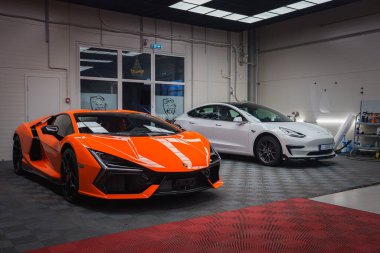 Canlı turuncu bir Lamborghini Revuelto ve beyaz bir Tesla Model 3 kareli zemini ve Tesla şarj istasyonu olan geniş, iyi aydınlatılmış bir garajda park halindeler..