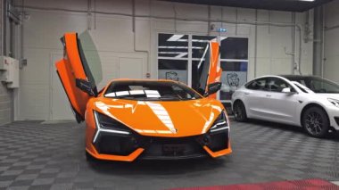 Canlı turuncu bir Lamborghini Revuelto, makas kapıları açık, modern bir galeride beyaz bir Tesla Model 3 'ün yanına park etmiş, endüstriyel tasarımlı..
