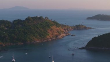 Tayland, Phuket Adası 'ndaki yeşil bir pelerin, demirli yelkenliler, küçük bir sahil ve altın ışık altında uzak bir ada..