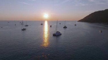 Gün batımında Tayland 'daki Phuket Körfezi' nin sakin hava manzarasında demirli yatlar, sakin sular ve solda ağaçlarla kaplı bir tepe yer alıyor..