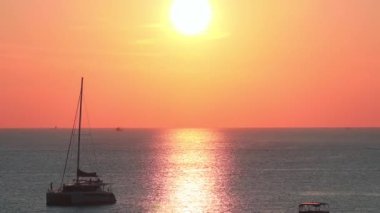 Phuket Adası, Tayland yakınlarındaki sakin okyanusun üzerinde sakin bir gün batımı, canlı gökyüzü, demirli yelkenliler ve daha büyük gemilerin uzak siluetleri..