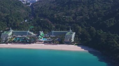 Tayland 'ın Phuket Adası' ndaki sakin bir sahil manzarasında turkuaz sular, yeşil çatıları olan beyaz bir otel ve yemyeşil orman tepeleri var..
