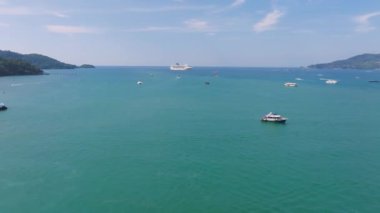 Tayland 'daki Phuket Adası' nın turkuaz suları, bir yolcu gemisi, küçük tekneler, deniz paraşütü, yemyeşil tepeler ve açık mavi bir gökyüzü..