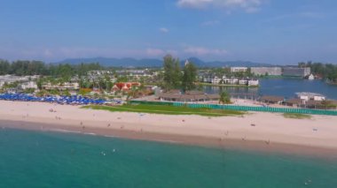 Tayland 'ın Phuket Adası' ndaki kumlu bir sahil manzarası, turkuaz sular, yemyeşil tepeler, güneşlenenler, şemsiyeler, botlar ve tatil köyleri..