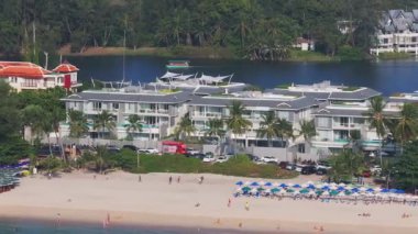 Tayland 'da canlı bir Phuket plajının havadan görünüşü, şemsiyeler, sahile gidenler, modern binalar, palmiye ağaçları ve geleneksel Tayland mimarisi yer alıyor..