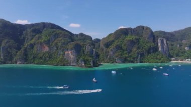 Tayland 'daki Phi Phi Adası' nın havadan görünüşü yeşil uçurumlar, turkuaz sular, botlar, kıyı şeridi yerleşimi ve açık mavi bir gökyüzünü gösteriyor..