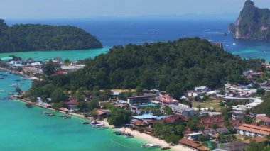 Tayland 'daki Phi Phi Adası' nın hava manzarasında turkuaz sular, yeşil tepeler, botlar ve renkli binalarla dolu canlı bir kıyı şeridi yer alıyor..