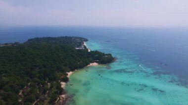 Tayland 'daki Phi Phi Adası' nın havadan görünüşü, yeşil tepeler, tropikal bitki örtüsü, küçük bir sahil, demirlemiş botlar ve geniş bir okyanus ufkunun yer aldığı.