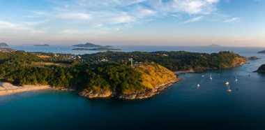 Tayland 'ın Phuket kentindeki Naiharn Sahili' nin havadan görünüşü, altın kumlar, turkuaz sular, demirlemiş yelkenliler, yemyeşil tepeler ve uzak bir deniz feneri içeriyor..