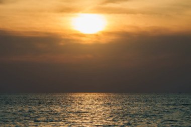 Okyanus üzerinde sakin bir günbatımı. Güneş kısmen bulutlar tarafından gizleniyor. Sıcak altın ışık, sakin denizi yansıtıyor. Bu da tropikal bir ortam olduğunu gösteriyor..