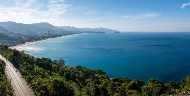 Aerial view of a sandy beach in Phuket, Thailand, bordered by lush greenery and clear blue sea. A winding road and rolling hills complete the scene. clipart