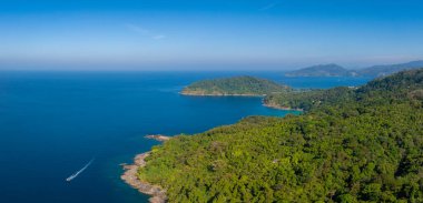 Tayland 'ın Phuket kentindeki tropikal kıyı şeridinin hava manzarası, yoğun yeşillik, canlı mavi bir deniz, hareketli bir tekne ve uzak orman tepeleri..