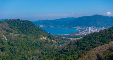 Phuket, Tayland 'ın havadan görünüşü, yemyeşil tepeler, canlı bir mavi koy, uzun beyaz bir bina ve çevreleyen dağlarla çevrili bir şehir manzarası..