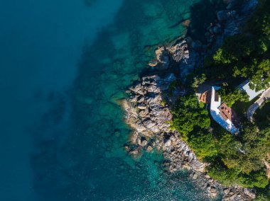 Tayland, Phuket 'te bir kıyı bölgesinin havadan görüntüsü. Turkuaz sular, kayalık kıyılar, yemyeşil yemyeşil ve kırmızı çatılı beyaz bir bina..